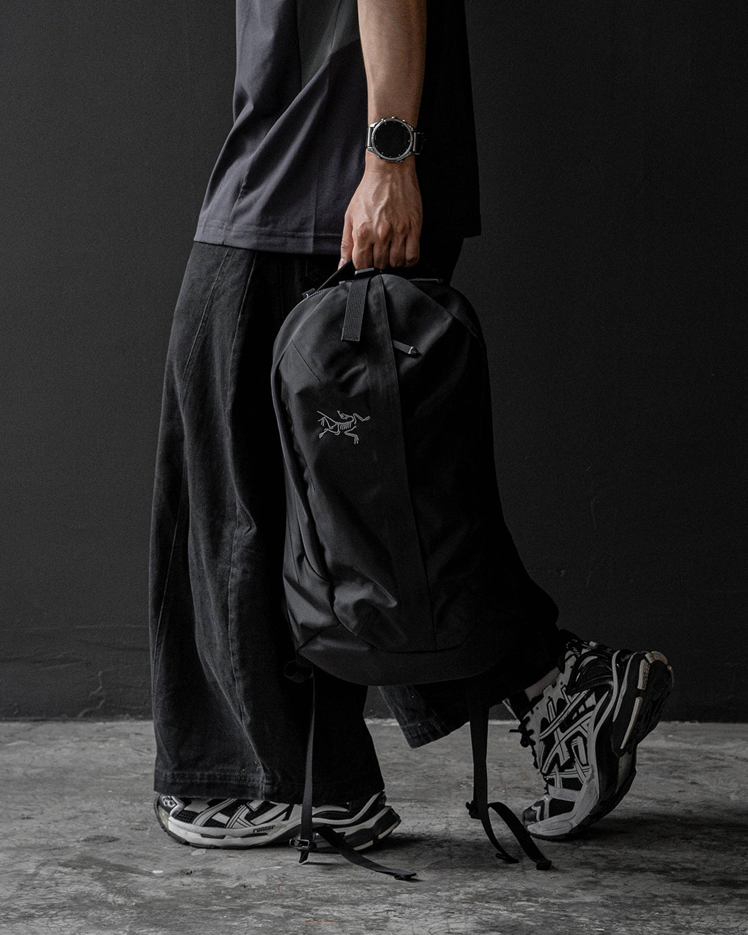 Arc’teryx Verro 20 Backpack สี Black ถือด้วยหูหิ้วด้านบน คู่กับยีนส์ดำและรองเท้าสีเงินดำ